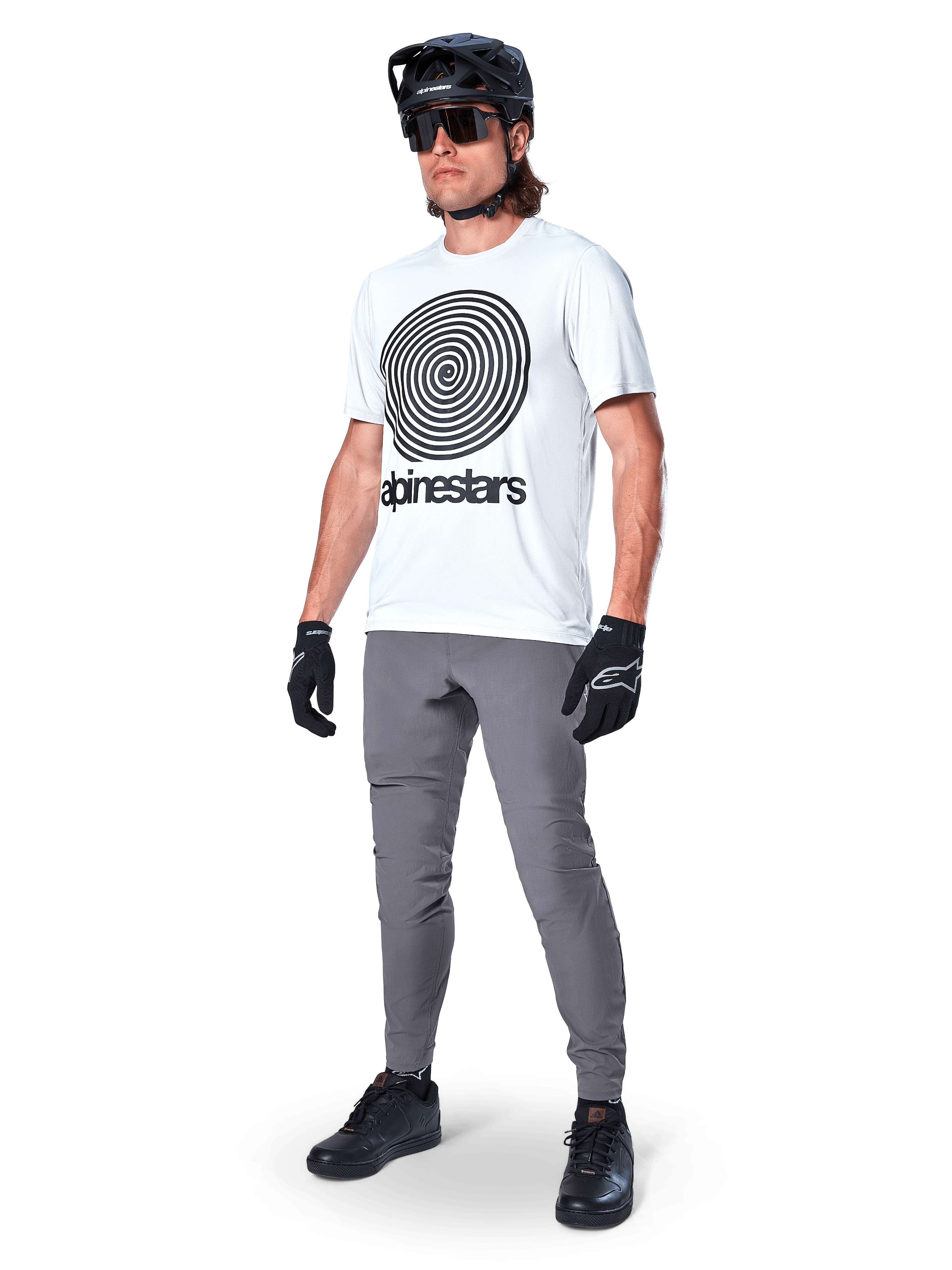 Alpinestars A-Dura Hose, MTB-Hose, Dunkelgrau, getragen mit weißem T-Shirt mit Spiralgrafik, schwarzem MTB-Helm, Sonnenbrille und schwarzen Handschuhen, strapazierfähiges 2-Wege-Stretch-Textil für den Offroad-Radsport