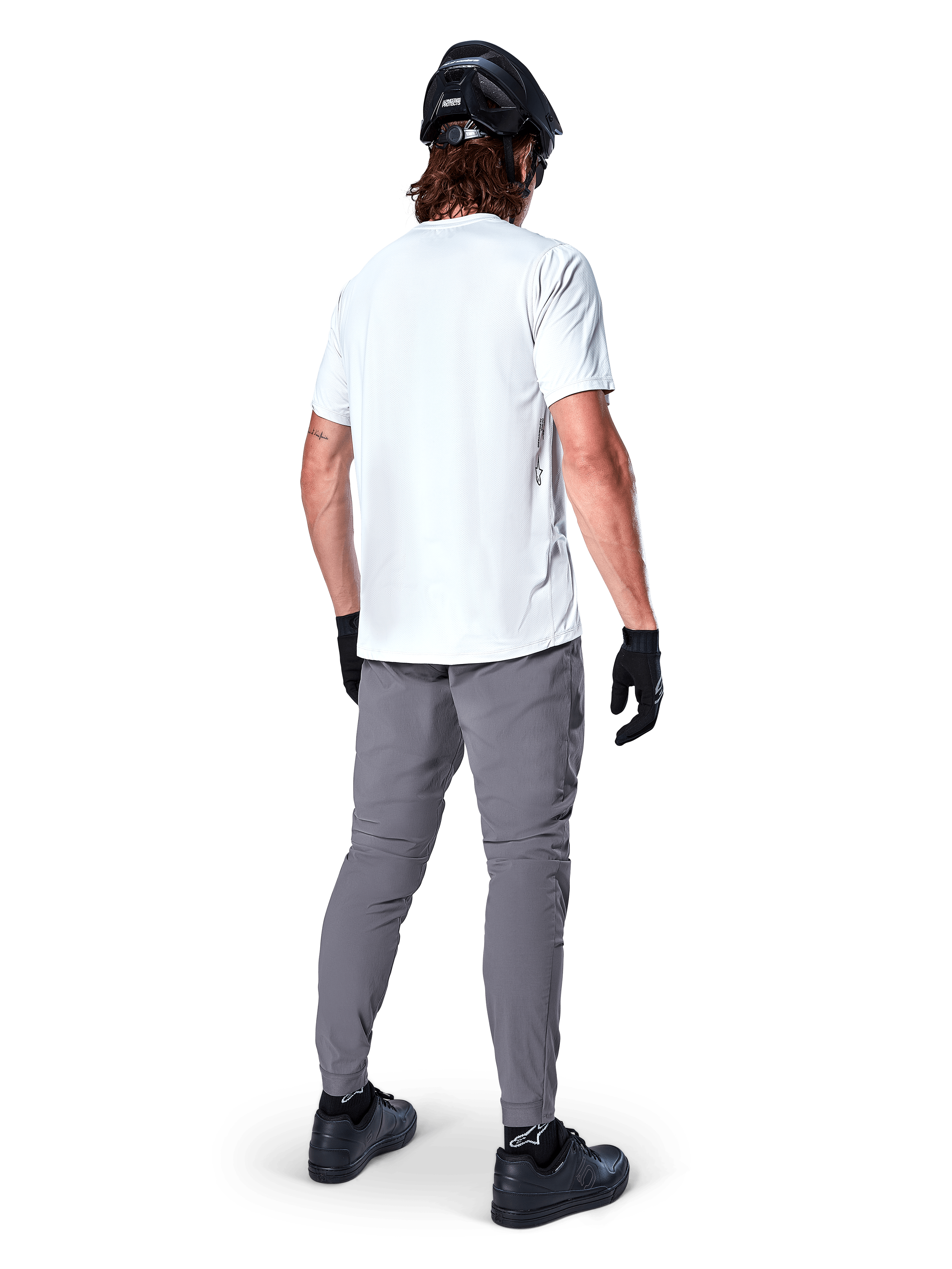 Alpinestars A-Dura Hose, dunkelgraue Mountainbike-Hose in der Rückansicht, getragen mit einem weißen Kurzarm-T-Shirt, schwarzem MTB-Helm, schwarzen Handschuhen und Schuhen, reguläre Passform mit elastischen Bündchen, strapazierfähige Offroad-Fahrradausrüstung