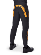 Pantalon Alpinestars A-Supra Race, pantalon de VTT noir, vue arrière avec un grand lettrage Alpinestars orange sur l'empiècement arrière, logo orange sur le bas de la jambe, coupe performance avec poche zippée, porté avec des chaussures noires
