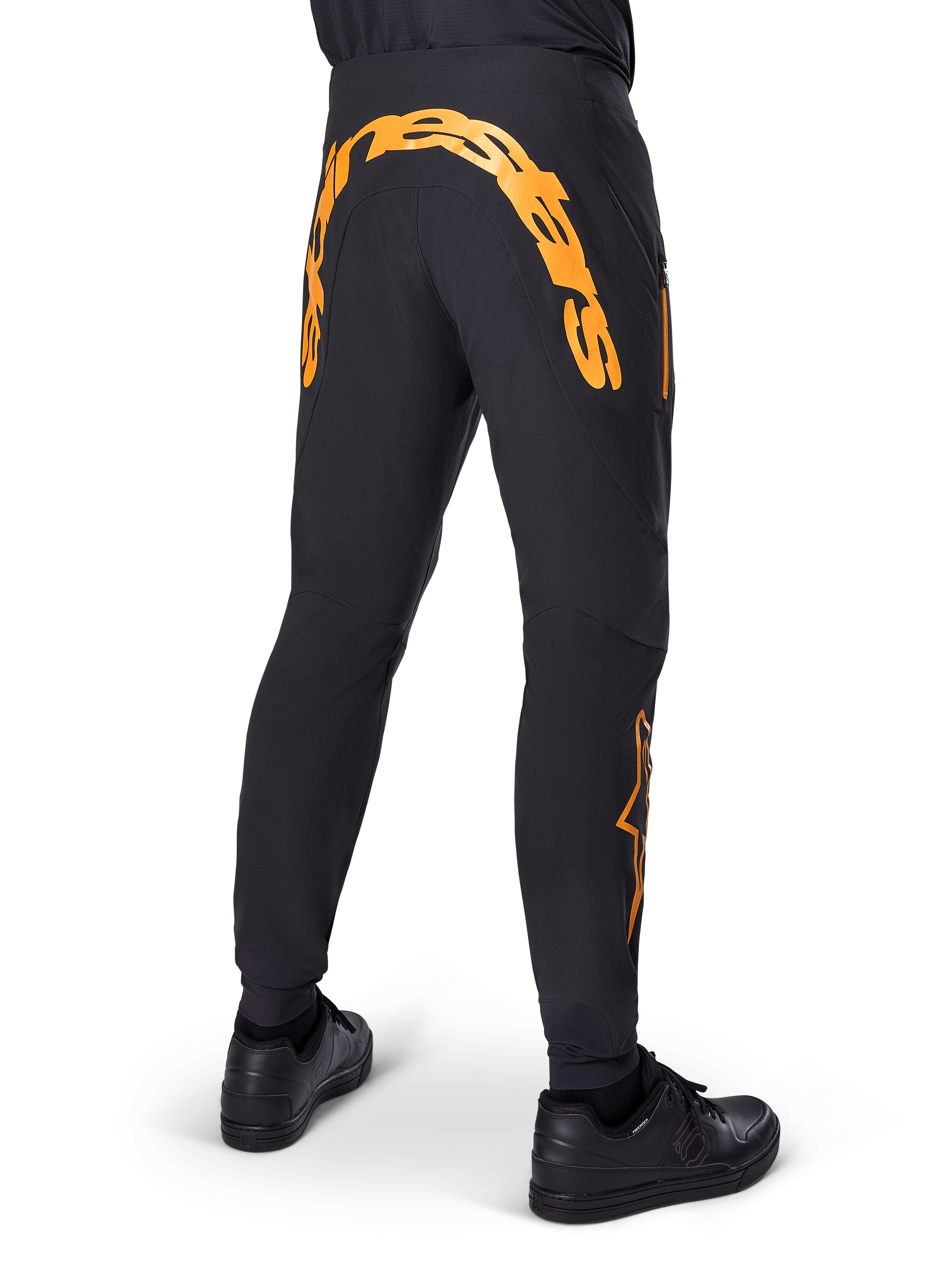 Pantalon Alpinestars A-Supra Race, pantalon de VTT noir, vue arrière avec un grand lettrage Alpinestars orange sur l'empiècement arrière, logo orange sur le bas de la jambe, coupe performance avec poche zippée, porté avec des chaussures noires