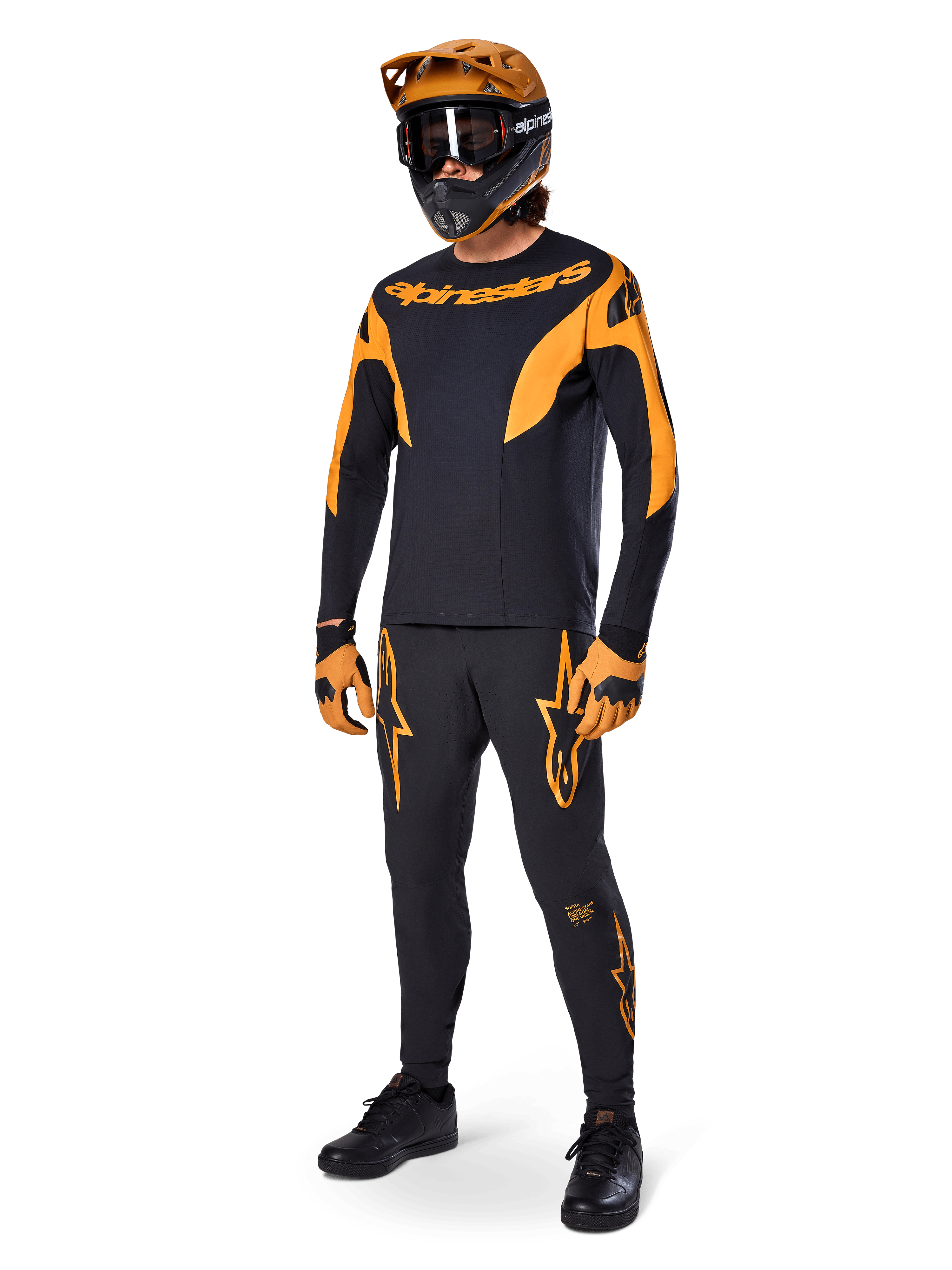 Pilote de VTT portant une tenue de course complète, pantalon de course Alpinestars A-Supra noir avec des accents orange, maillot à manches longues noir et orange assorti, casque orange avec lunettes noires, gants orange et noir, et chaussures noires, design de kit de course VTT professionnel