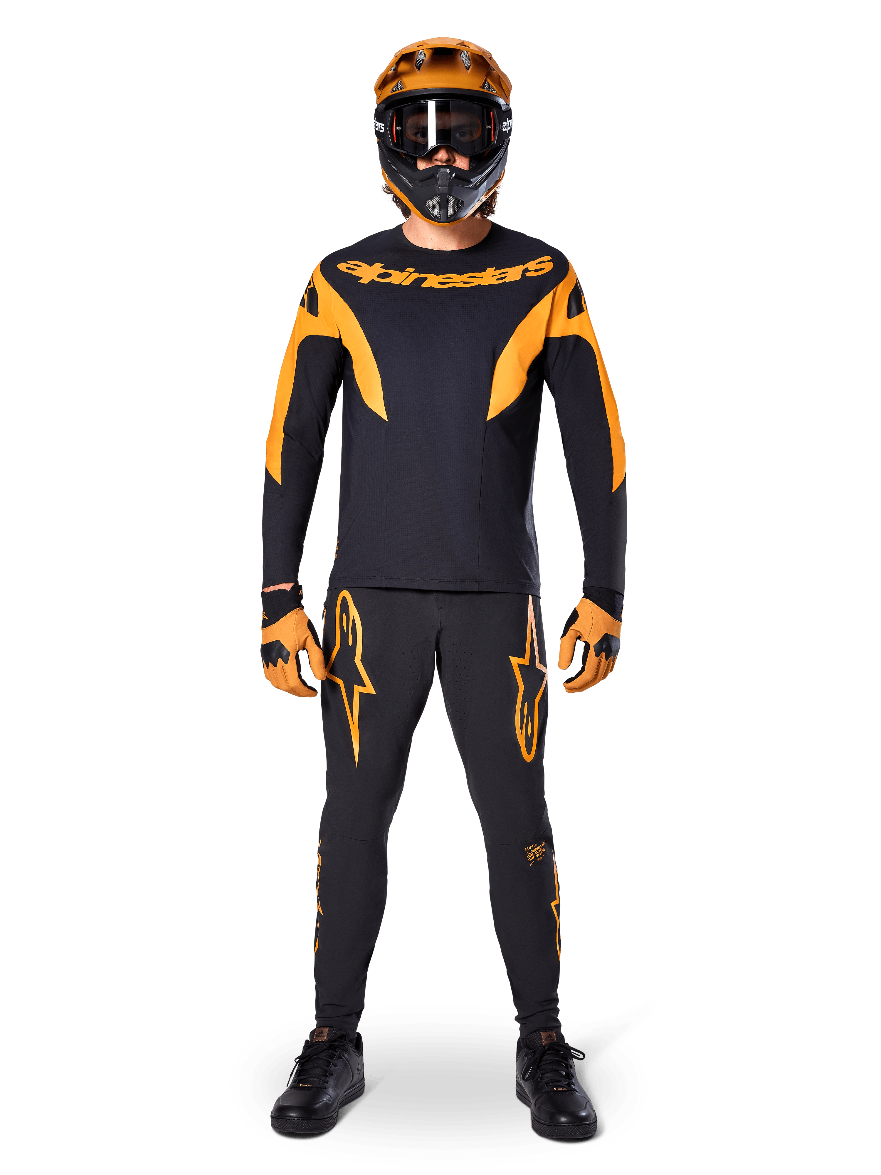 Alpinestars A-Supra Race Pants, pantalon de course VTT, noir et orange, porté par un pilote avec une tenue complète comprenant un maillot noir et orange, un casque orange et des gants assortis, avec logos étoiles orange sur les jambes