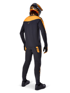 Alpinestars A-Supra Race Pants, pantalon de course VTT, noir avec logos orange, vue de dos sur un pilote portant un maillot, un casque et des gants noir et orange assortis, mettant en avant le logo inspiré du MX sur l'empiècement arrière et la coupe performance pour les sentiers techniques.