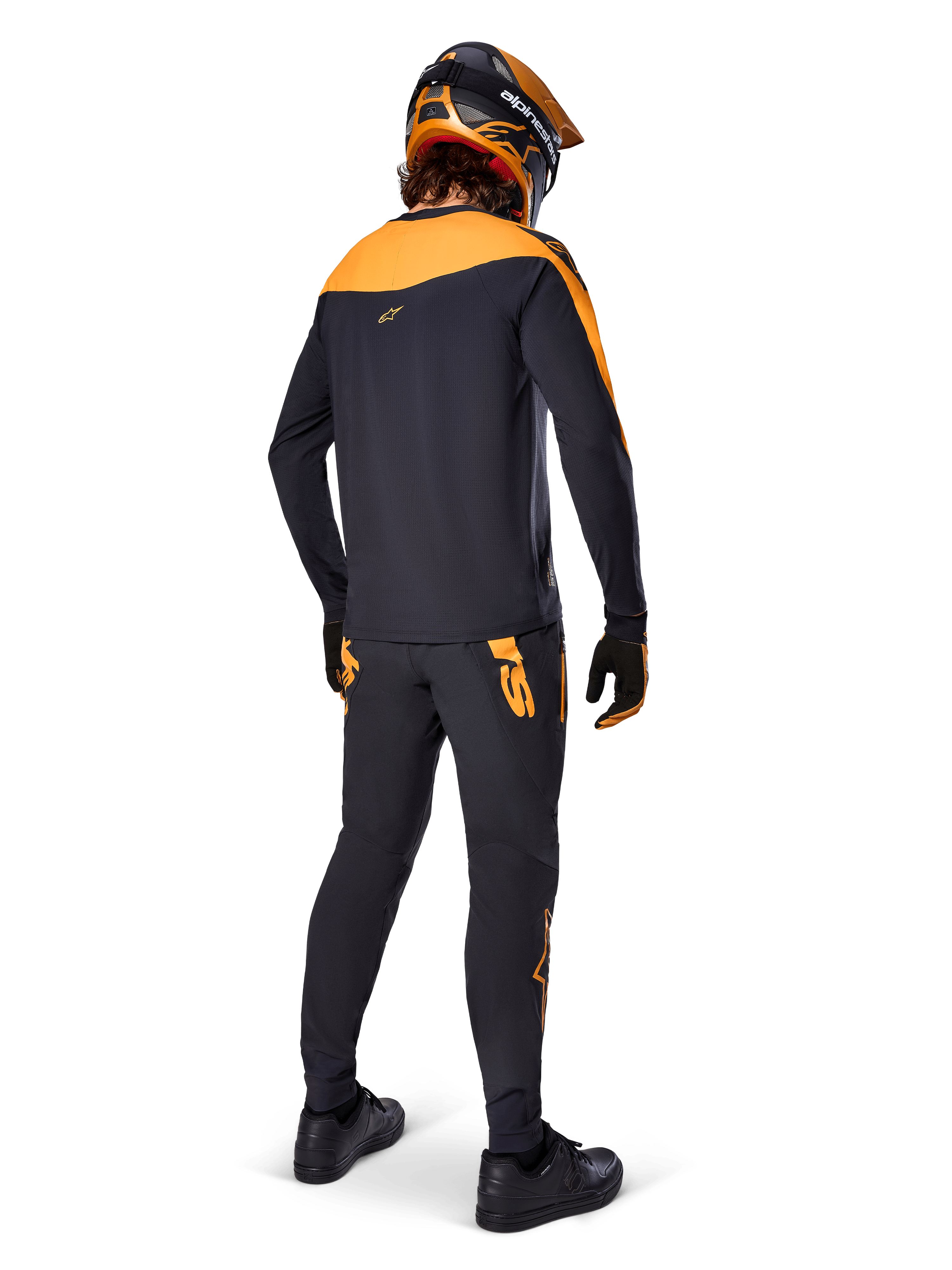 Alpinestars A-Supra Race Pants, pantalon de course VTT, noir avec logos orange, vue de dos sur un pilote portant un maillot, un casque et des gants noir et orange assortis, mettant en avant le logo inspiré du MX sur l'empiècement arrière et la coupe performance pour les sentiers techniques.