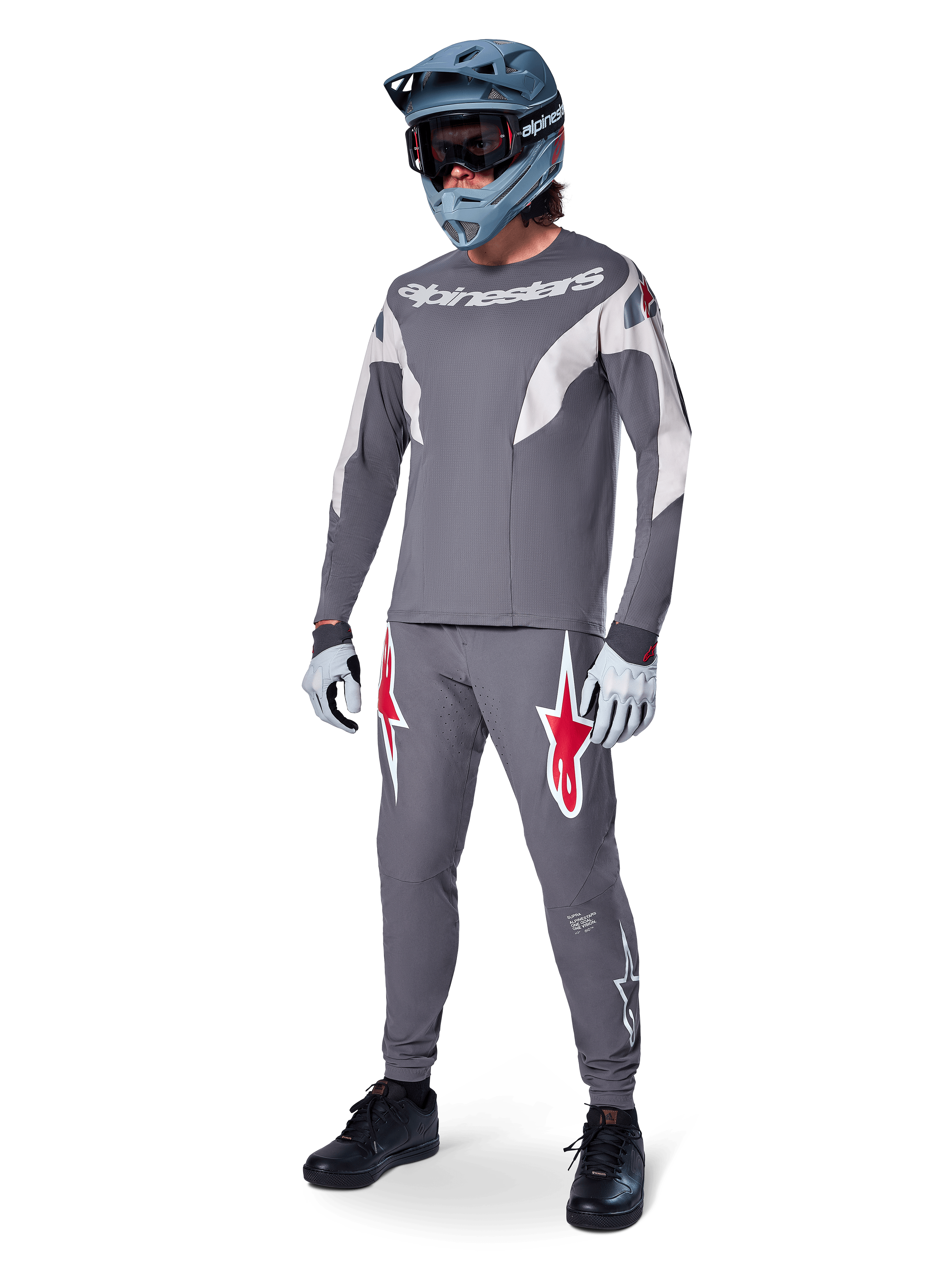 Pantalon Alpinestars A-Supra Race en gris foncé, avec des logos rouges et blancs sur les cuisses, porté par un pilote avec un maillot gris assorti, un casque intégral bleu-gris et des gants gris clair, équipement haute performance pour la course VTT