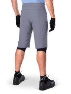 Short Alpinestars A-Dura, short de VTT gris foncé, vue arrière portée par un cycliste avec gants noirs, genouillères noires et chaussettes noires à logo Alpinestars, construction durable pour le cyclisme tout-terrain