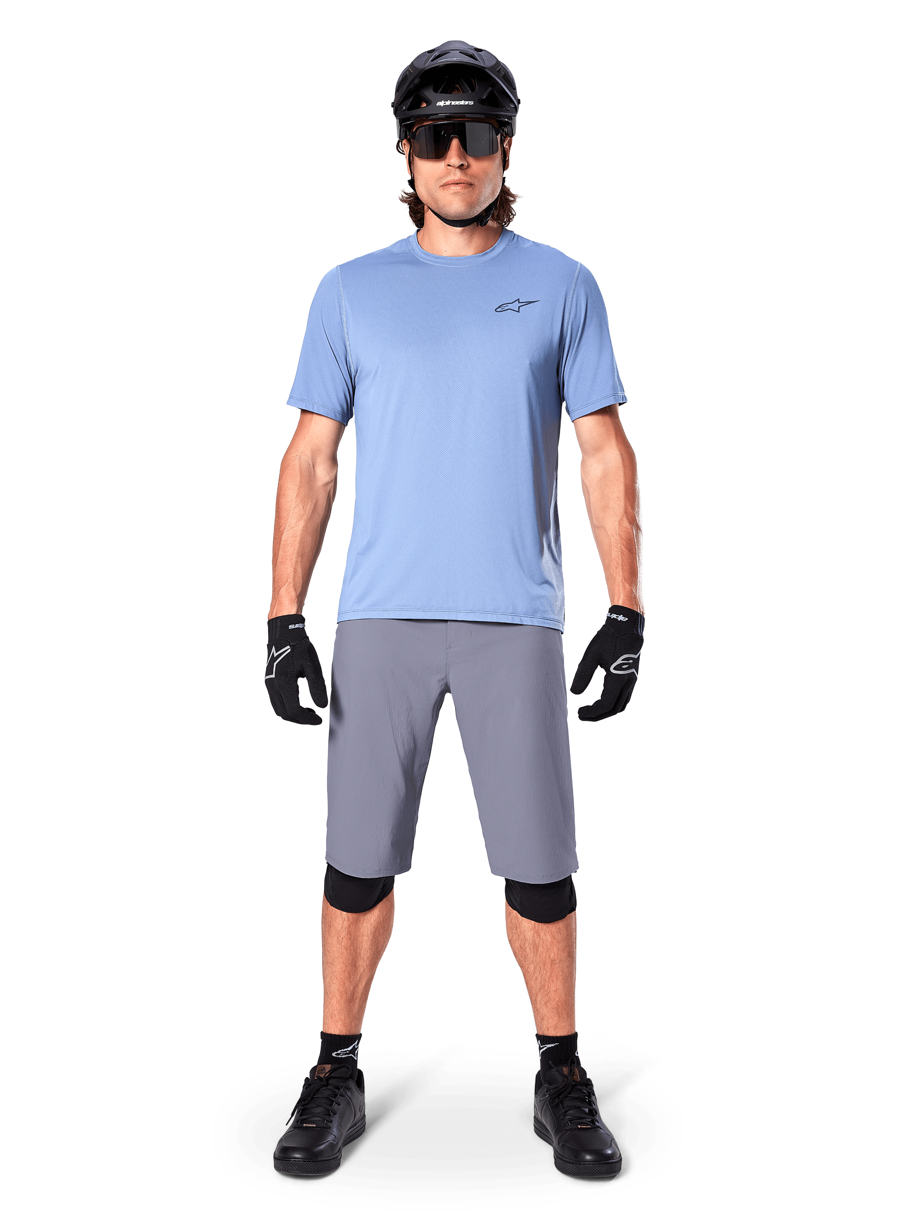 Cycliste portant un short Alpinestars A-Dura gris foncé, avec un t-shirt à manches courtes bleu clair, un casque noir, des lunettes de soleil et des genouillères noires, tenue de VTT complète pour la performance tout-terrain.