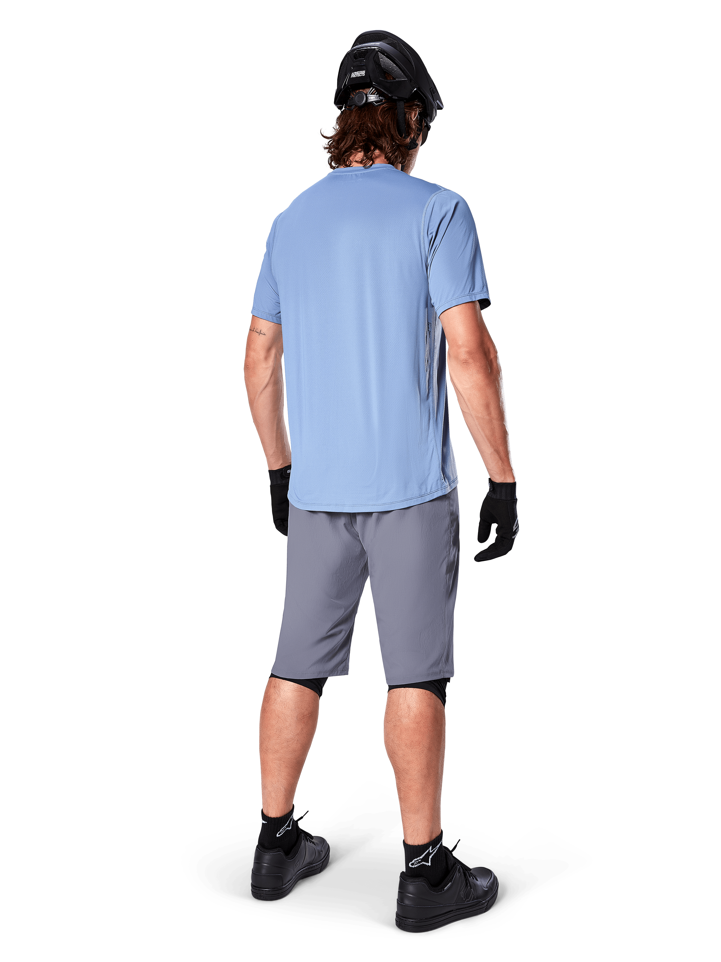 Alpinestars A-Dura Shorts, short de VTT gris foncé, vue de dos, porté avec un maillot bleu clair à manches courtes et un casque de VTT noir, gants et chaussettes noirs avec chaussures noires, équipement fonctionnel et durable pour le cyclisme tout-terrain