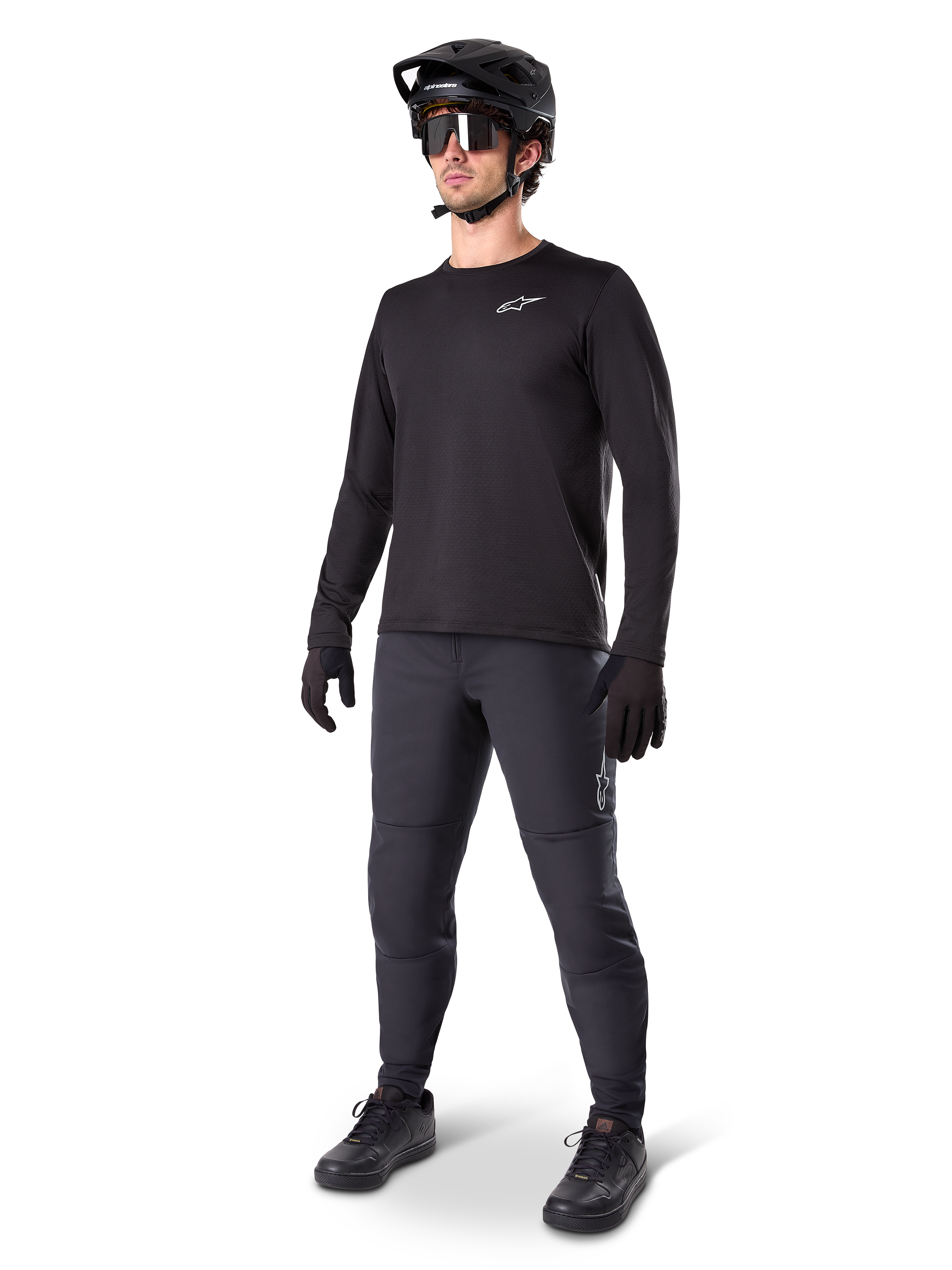 Modèle portant le maillot thermique Alpinestars A-Dura, maillot de VTT noir à manches longues, casque de VTT noir, lunettes de soleil et pantalon gris foncé, avec un petit logo blanc sur la poitrine, conception en polaire isolante pour rouler par temps froid
