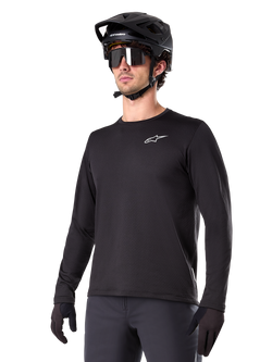 Alpinestars A-Dura Thermal Jersey, schwarzes Langarm-Mountainbike-Trikot, weißes Brust-Logo, getragen mit schwarzem Helm und Handschuhen, schlichtes sportliches Design für Trail-Fahrten