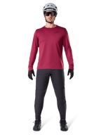 Modèle masculin portant le maillot thermique Alpinestars A-Dura bordeaux, haut de cyclisme isolé à manches longues, associé à un pantalon de VTT gris foncé, un casque blanc, des lunettes de soleil noires et des gants noirs, équipement professionnel de VTT pour temps froid