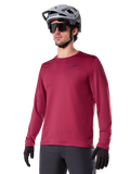 Maillot thermique Alpinestars A-Dura, maillot de VTT à manches longues, rouge bordeaux, avec doublure en polaire isolante et petit logo noir sur la poitrine, porté avec un casque gris, des lunettes de soleil noires et des gants noirs, conçu pour le cyclisme par temps froid