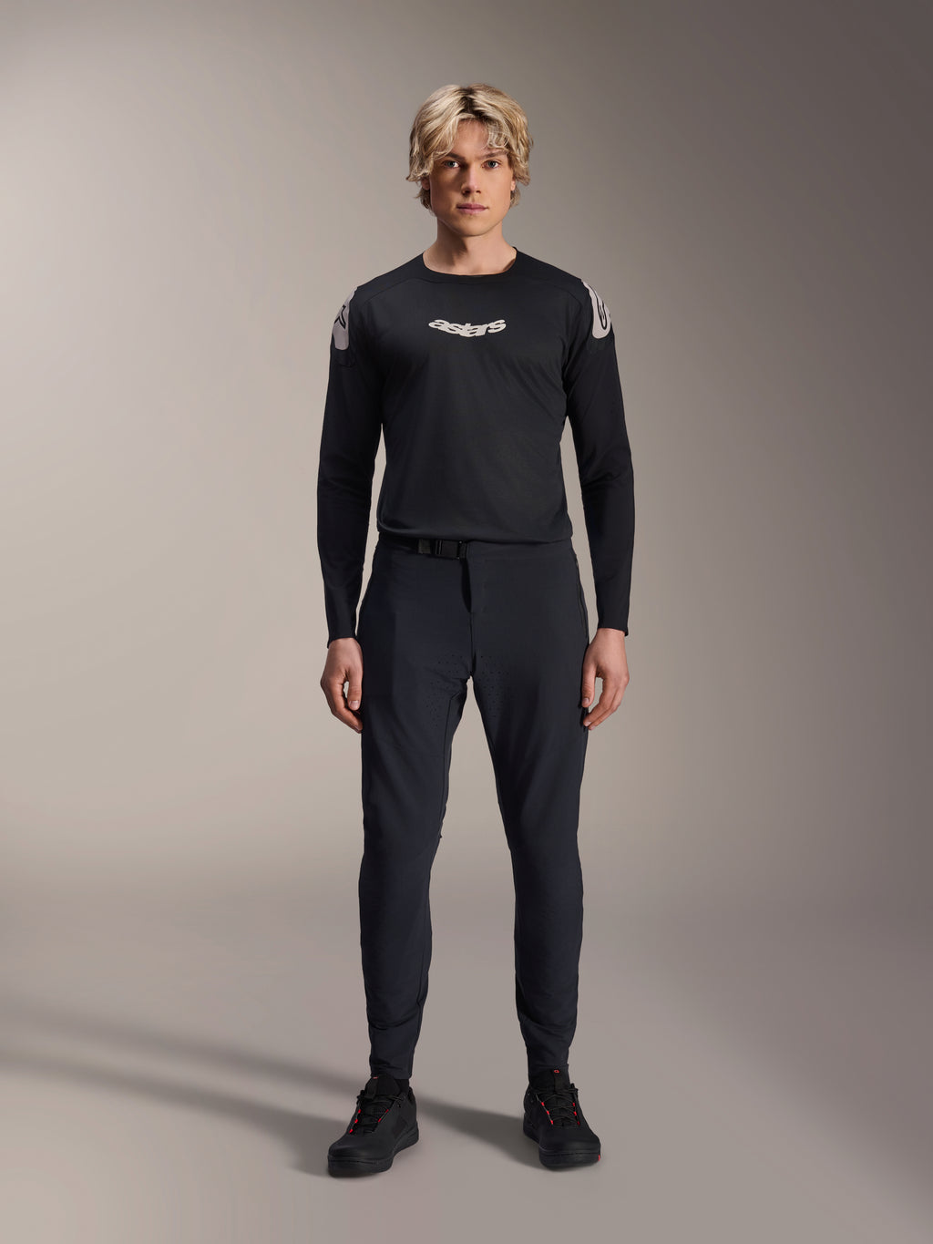 Alpinestars A-Aria Elite Pants, Black Titanium MTB-broek, gedragen door een mannelijk model met een bijpassende zwarte jersey met lange mouwen en sneakers, slim-fit prestatiegericht ontwerp met magnetische gespsluiting, technische mountainbikekleding