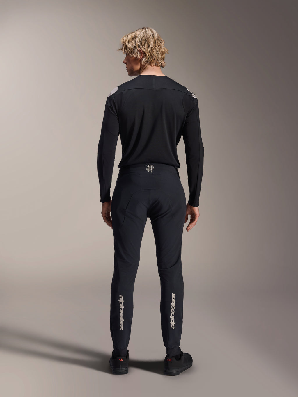Alpinestars A-Aria Elite broek, Black Titanium, achteraanzicht op mannelijk model, met zilveren Alpinestars-branding op de onderbenen, technische MTB-broek met prestatiepasvorm en vochtafvoerend detail in de tailleband