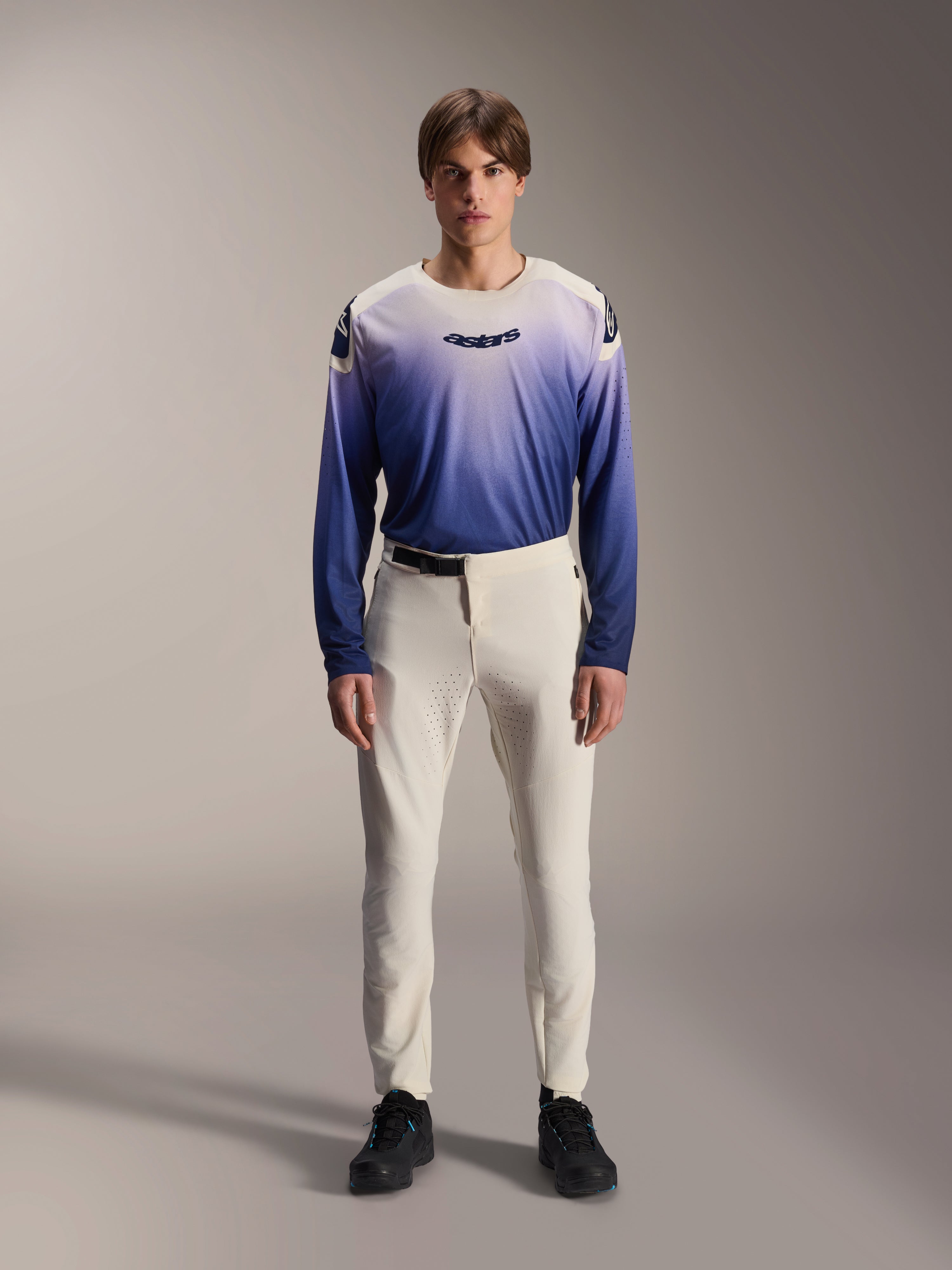 Alpinestars A-Aria Elite broek, ecru prestatiegerichte MTB-broek, gebroken witte fietsbroek met laser-geperforeerde dijpanelen en magnetische gesp, gedragen met blauw verloopshirt met lange mouwen en zwarte schoenen, prestatiepasvorm voor trailrijden