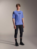 Mannelijk model in een zwarte Alpinestars A-Aria Lite Short, gecombineerd met een blauwe A-Aria Polartec Elite Jersey met korte mouwen, zwarte kniebeschermers en zwarte fietsschoenen, technische mountainbike-kleding voor trailrijden.