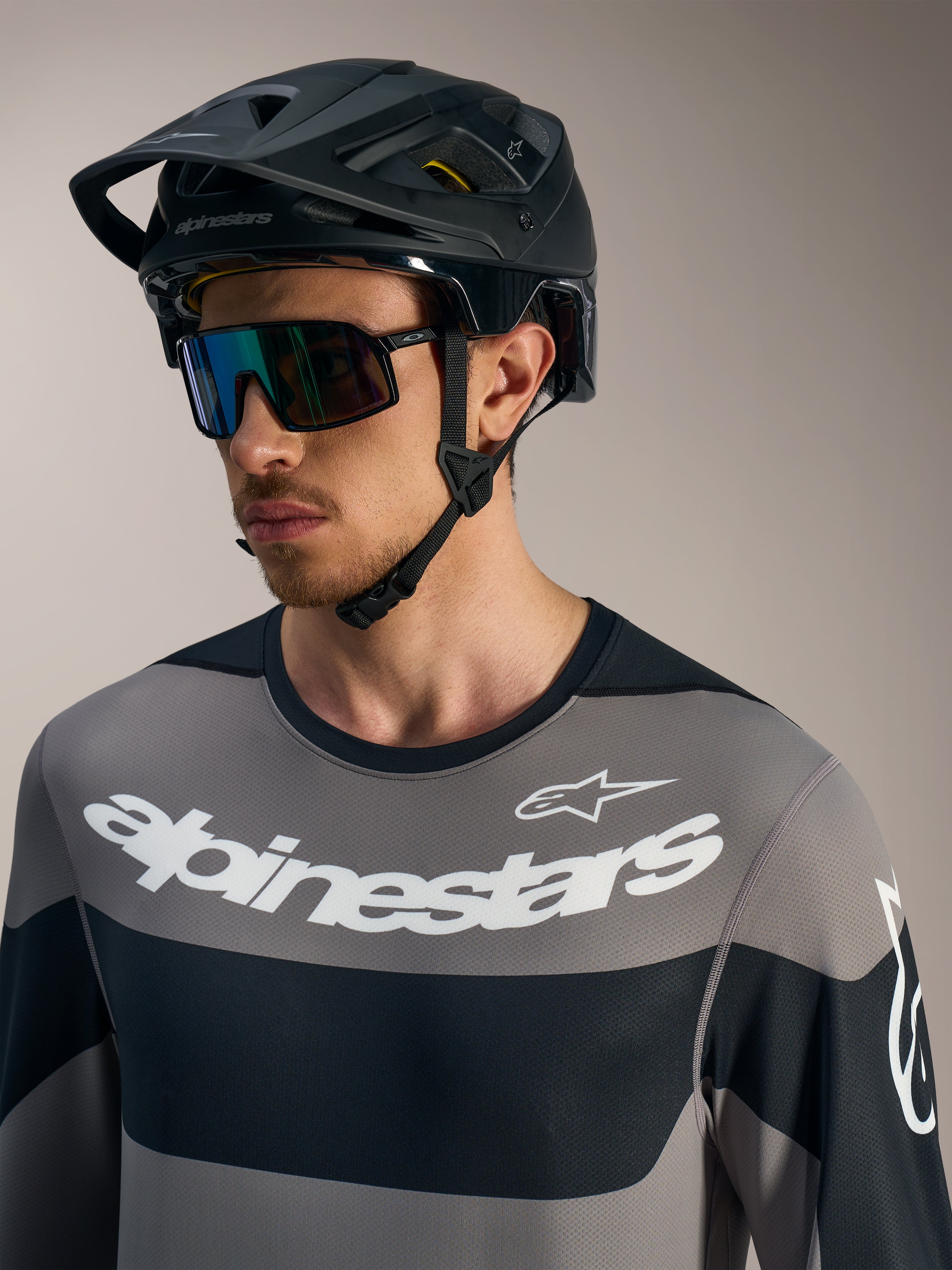 Alpinestars A-Dura Race jersey met lange mouwen in grijs, witte logo's, zwarte MTB-helm en sportieve zonnebril.