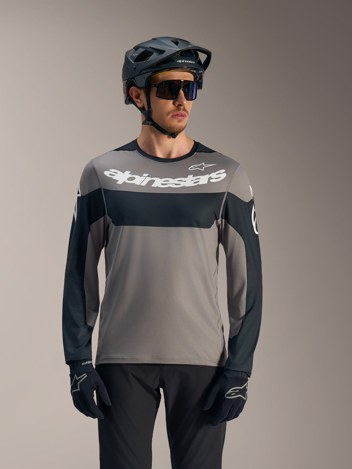 Grijs-zwarte A-Dura Race jersey met lange mouwen, wit Alpinestars-logo en MTB-helm voor trailrijden.