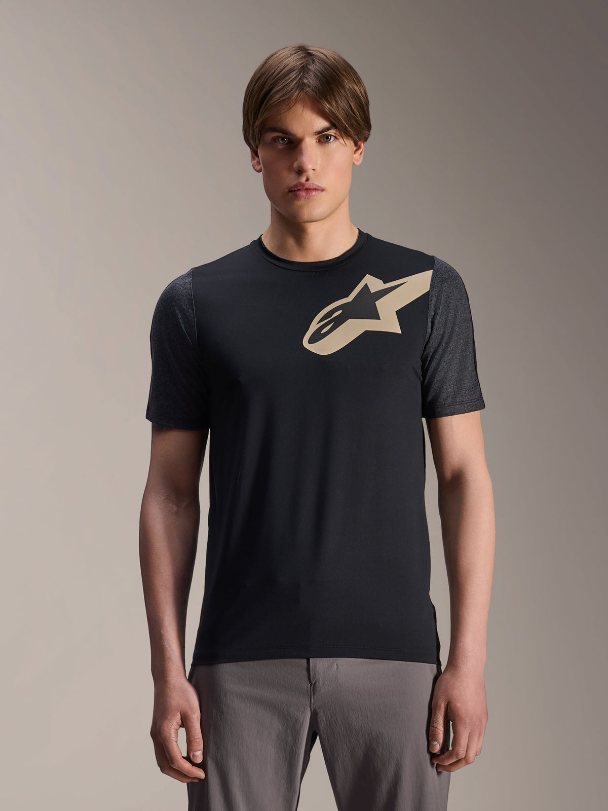 Mannequin homme portant le maillot à manches courtes Alpinestars A-Dura Lipan, maillot de VTT noir avec logo étoile beige et manches gris foncé, associé à un pantalon de cyclisme gris, tenue de performance décontractée
