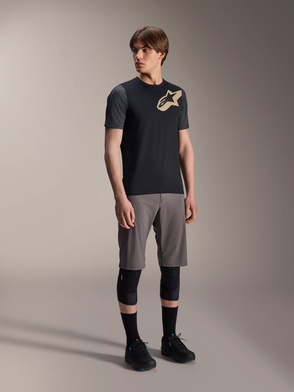 Männliches Model im Alpinestars A-Dura Lipan Kurzarmtrikot in Schwarz, mit dunkelgrauen Ärmeln und einem hellbraunen Astars-Logo auf der Brust, kombiniert mit grauen Mountainbike-Shorts, schwarzen Knieschonern und schwarzen Radschuhen, abgestimmte Mountainbike-Fahrausrüstung