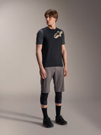 Männliches Model im Alpinestars A-Dura Lipan Kurzarmtrikot in Schwarz, mit dunkelgrauen Ärmeln und einem hellbraunen Astars-Logo auf der Brust, kombiniert mit grauen Mountainbike-Shorts, schwarzen Knieschonern und schwarzen Radschuhen, abgestimmte Mountainbike-Fahrausrüstung