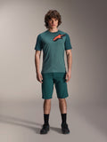 Alpinestars A-Dura Shorts in Dark Pine, mountainbike-short voor heren, getoond op een model met een bijpassende petrolkleurige jersey met korte mouwen met een rood en zwart sterlogo, zwarte sokken en fietsschoenen, casual sportieve mountainbike-kleding