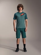 Alpinestars A-Dura Shorts in Dark Pine, mountainbike-short voor heren, getoond op een model met een bijpassende petrolkleurige jersey met korte mouwen met een rood en zwart sterlogo, zwarte sokken en fietsschoenen, casual sportieve mountainbike-kleding