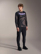 Modèle masculin portant le maillot à manches longues Alpinestars A-Dura Hollow Camo, motif Black Camo avec graphisme 'astars' violet sur la poitrine, associé à un pantalon technique noir et des chaussures noires, équipement de VTT décontracté