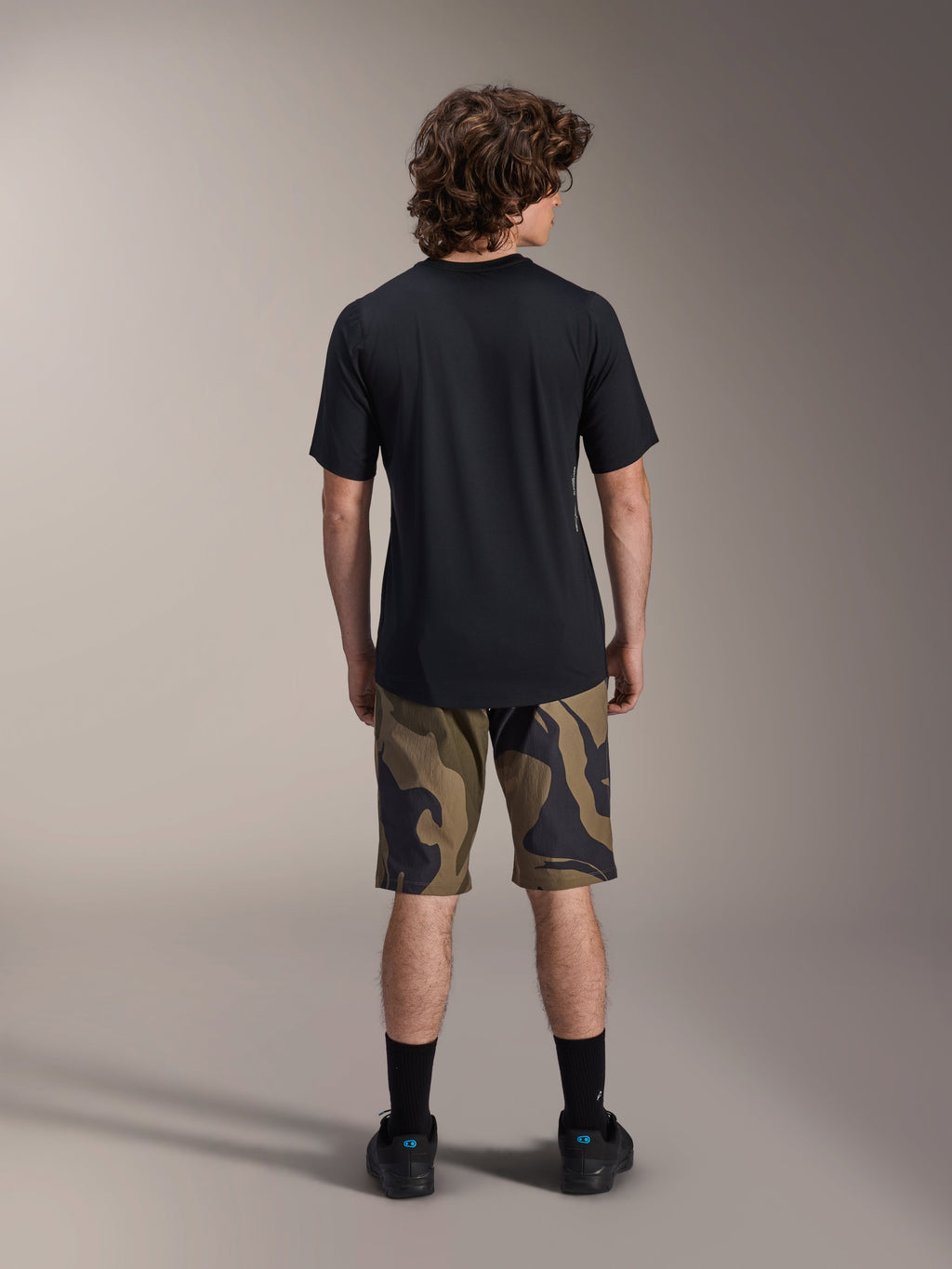 Alpinestars A-Dura Astar Kurzarm-Trikot, Schwarz, Rückansicht an einem Modell, kombiniert mit MTB-Shorts mit Camouflage-Muster und schwarzem Schuhwerk, lässige technische Mountainbike-Bekleidung