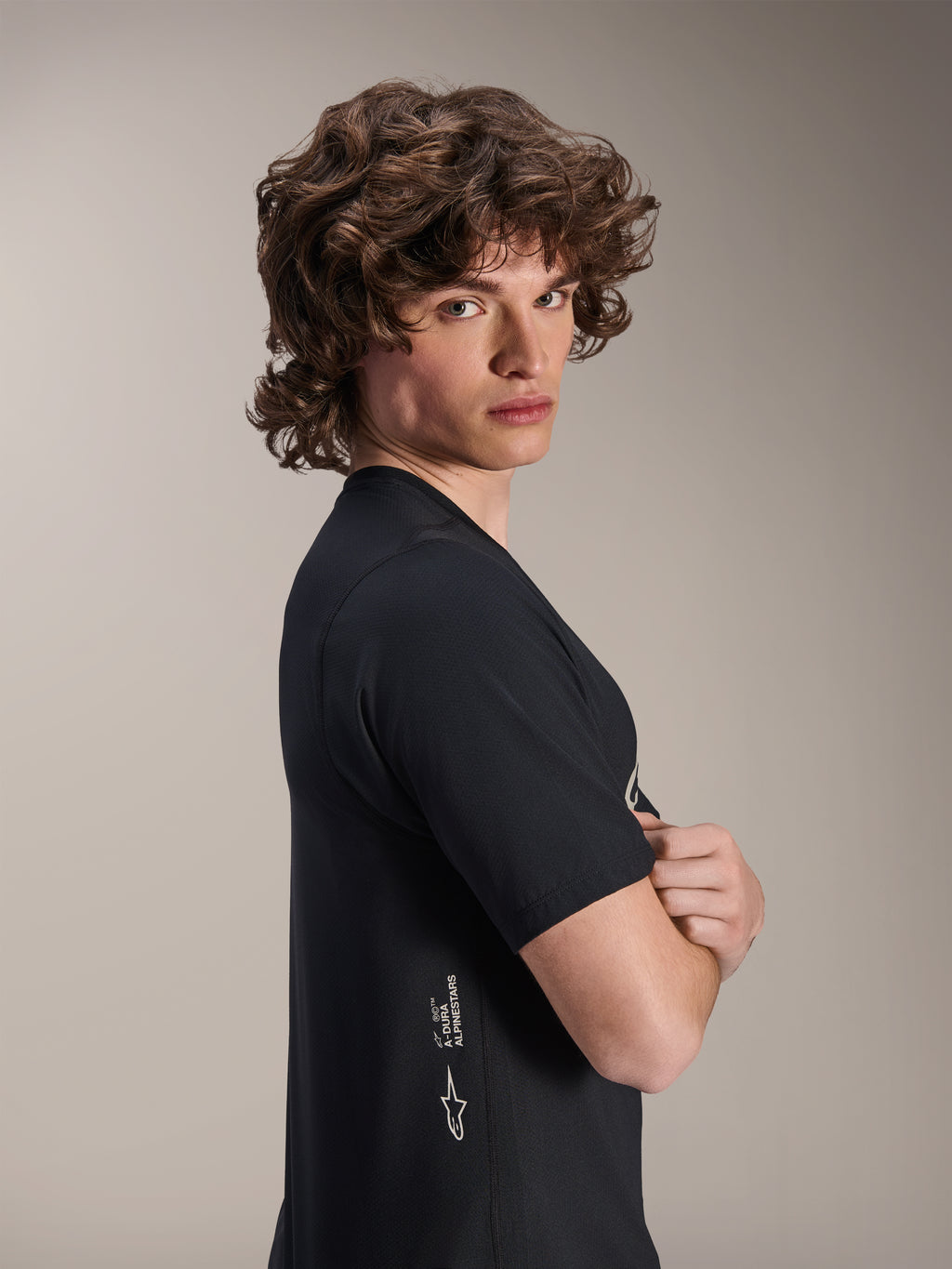 Alpinestars A-Dura Astar Jersey, Kurzarm-MTB-Trikot, Schwarz, männliches Model posiert im Profil mit verschränkten Armen, mit weißem Alpinestars-Branding an der unteren Seite, lässiges sportliches Design für Mountainbiking.