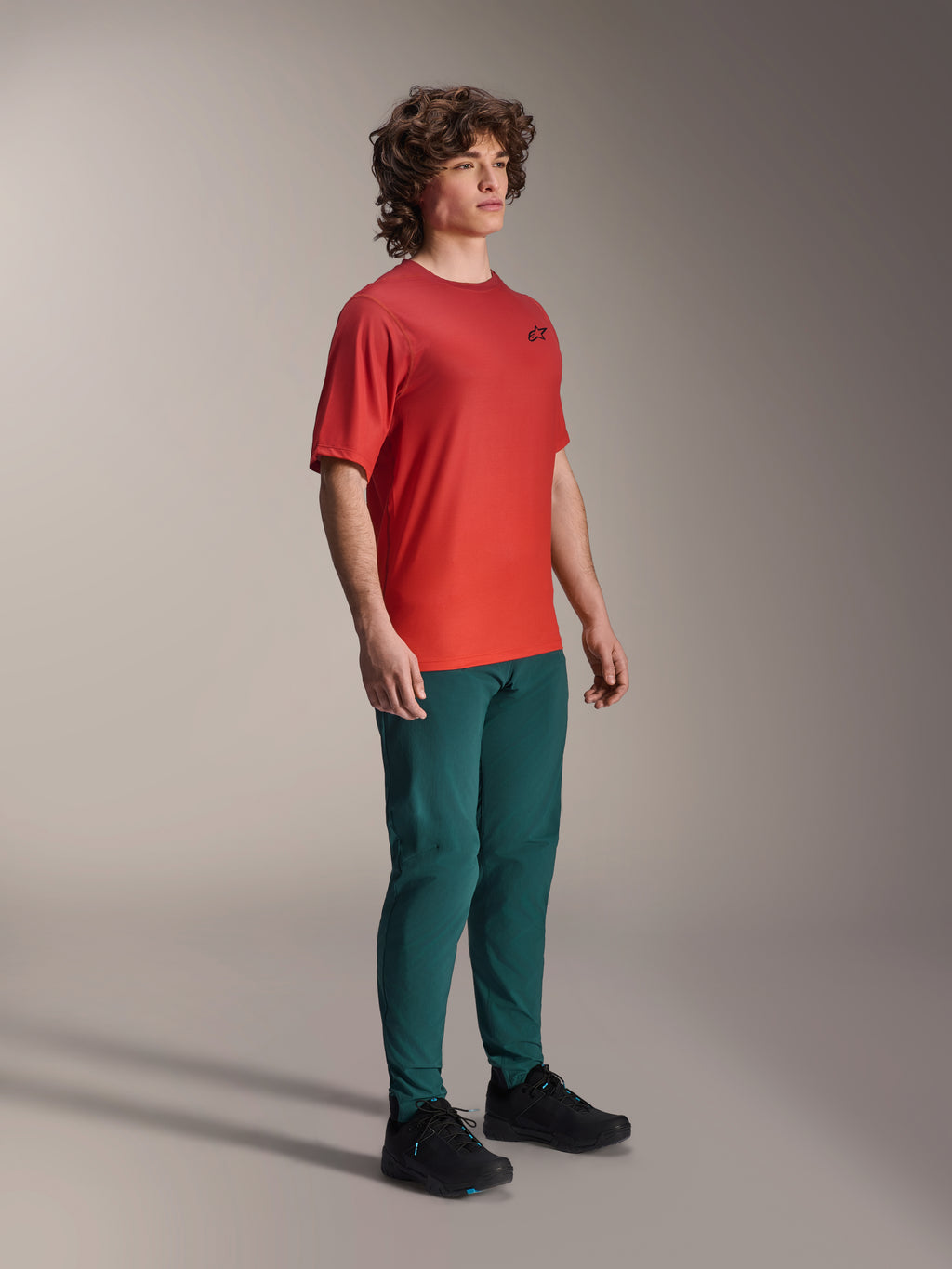Model trägt Alpinestars A-Dura Hose in Dark Pine, kombiniert mit einem roten A-Dura Rise Kurzarmtrikot und schwarzen MTB-Schuhen, sportliche Mountainbike-Bekleidung in einer Studio-Umgebung