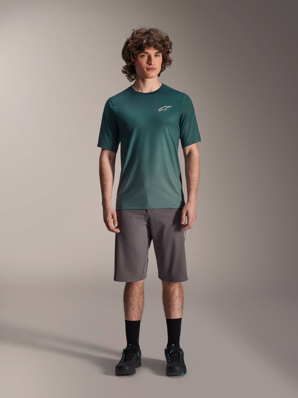 Mannelijk model draagt Alpinestars A-Dura Rise Jersey - Korte Mouwen in Silver Pine, groen fietsshirt met klein wit logo op de borst, gecombineerd met een grijze mountainbike short en zwart schoeisel, casual MTB-trailuitrusting