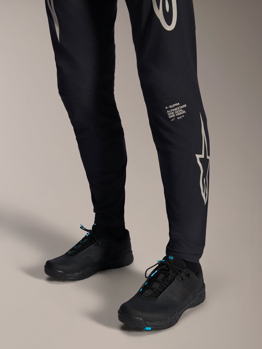 Pantalon Alpinestars A-Supra Melt, pantalon de VTT noir, avec logo et lettrage Alpinestars blancs sur la jambe, porté avec des chaussures de cyclisme noires aux accents bleus, équipement tout-terrain haute performance