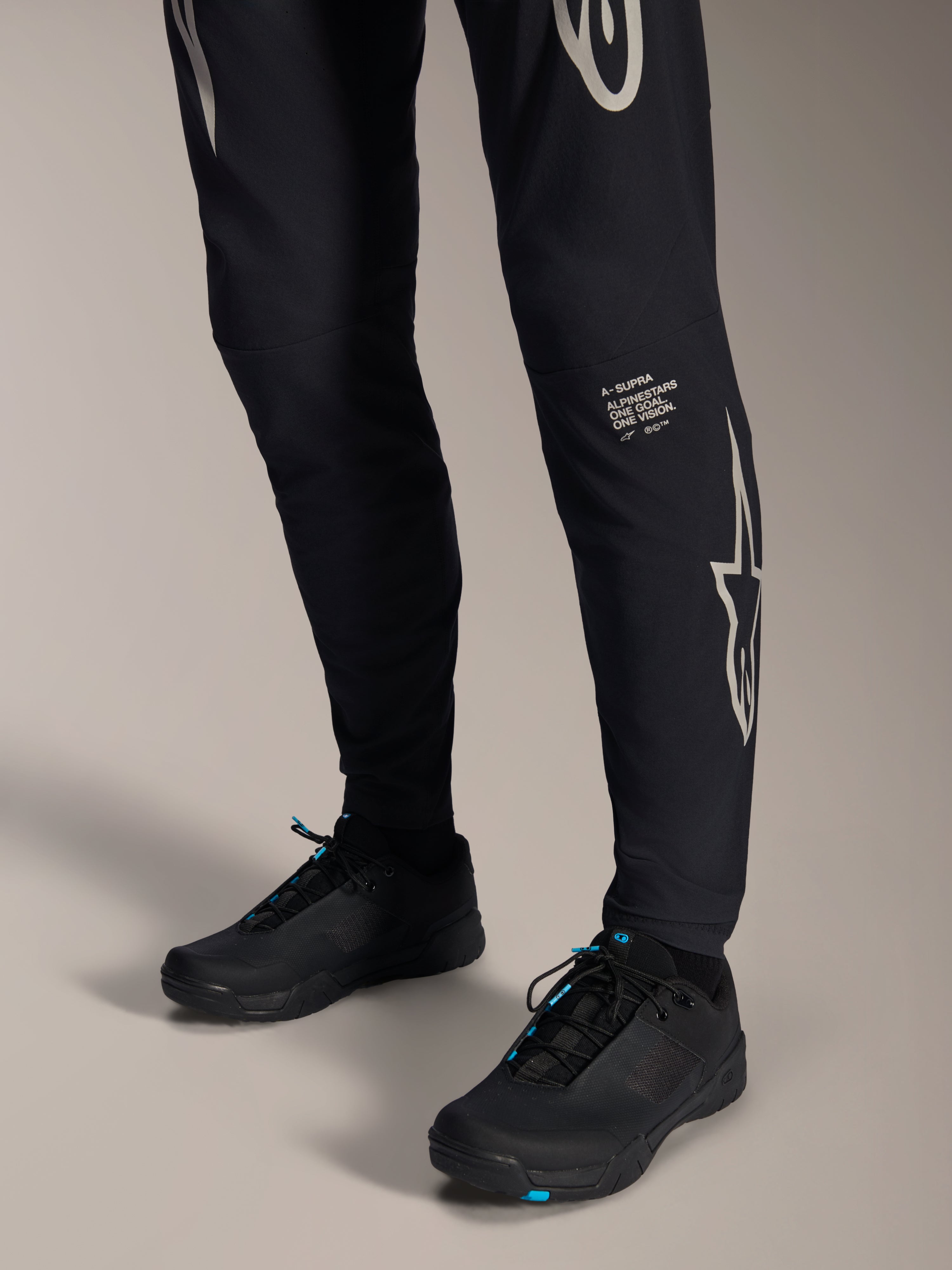 Pantalon Alpinestars A-Supra Melt, pantalon de VTT noir, avec logo et lettrage Alpinestars blancs sur la jambe, porté avec des chaussures de cyclisme noires aux accents bleus, équipement tout-terrain haute performance