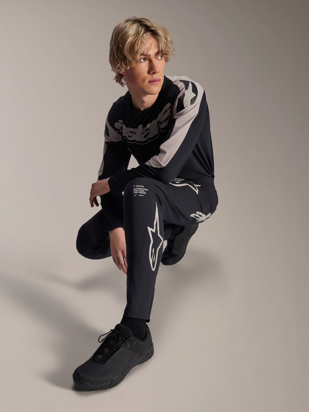 Pantalon et maillot à manches longues Alpinestars A-Supra Melt, noir et gris, avec logos blancs et logos Astars audacieux, portés par un mannequin masculin dans une pose accroupie, tenue de course VTT coordonnée