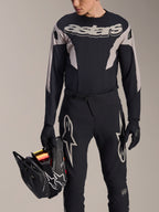 Maillot à manches longues Alpinestars A-Supra Melt, noir avec empiècements graphiques gris et marquage 'astars' sur la poitrine, porté avec un pantalon de VTT et des gants noirs tout en tenant un casque intégral noir, tenue de course VTT haute performance