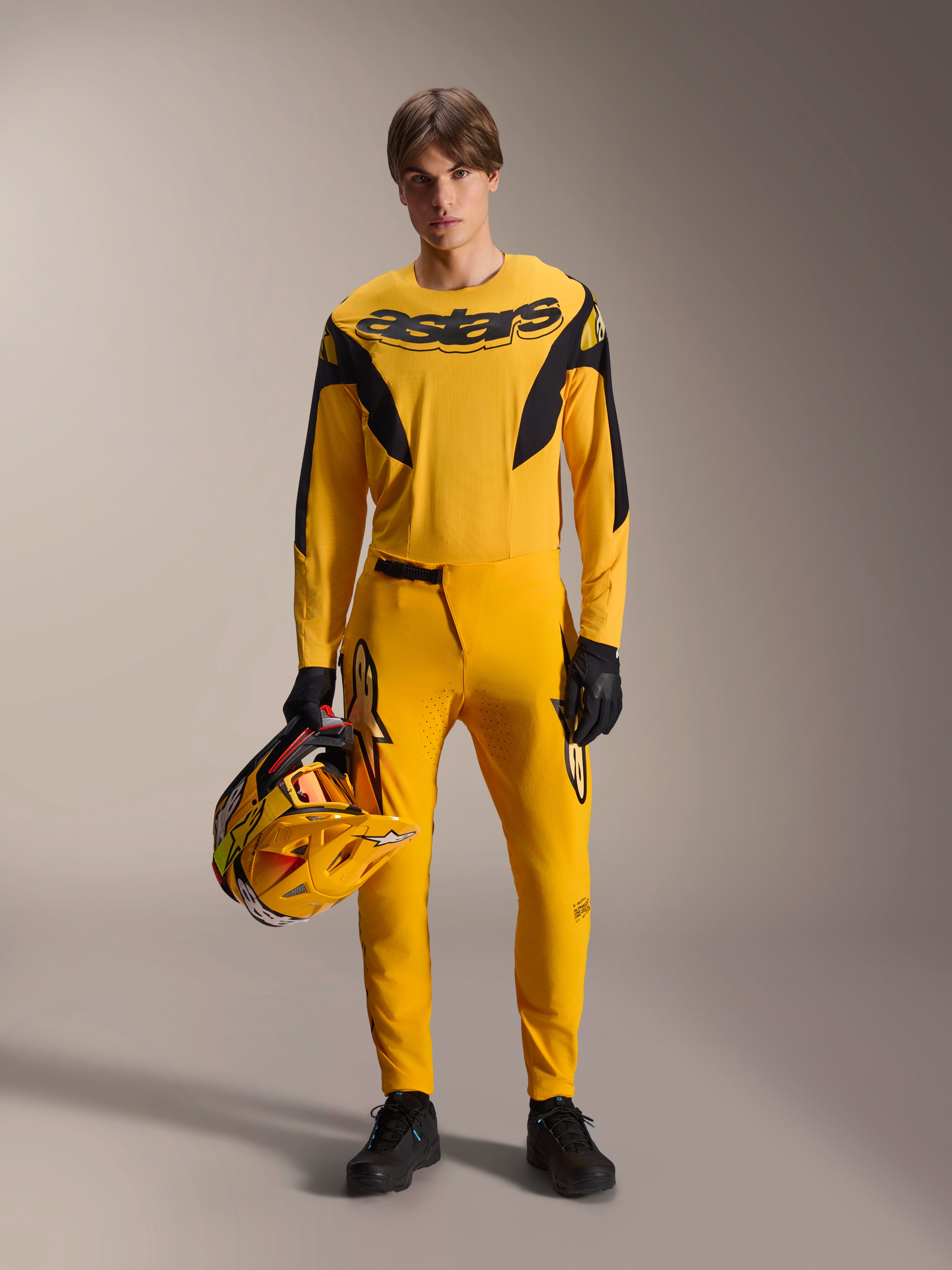 Mannelijk model in de Limited Edition A-Supra Melt Gold Gear-Set van Alpinestars, donkergeel jersey met lange mouwen en zwart Astars-logo, bijpassende gele broek met zwarte accenten, houdt een gele en zwarte helm vast, zwarte handschoenen en technische schoenen, mountainbike race-outfit
