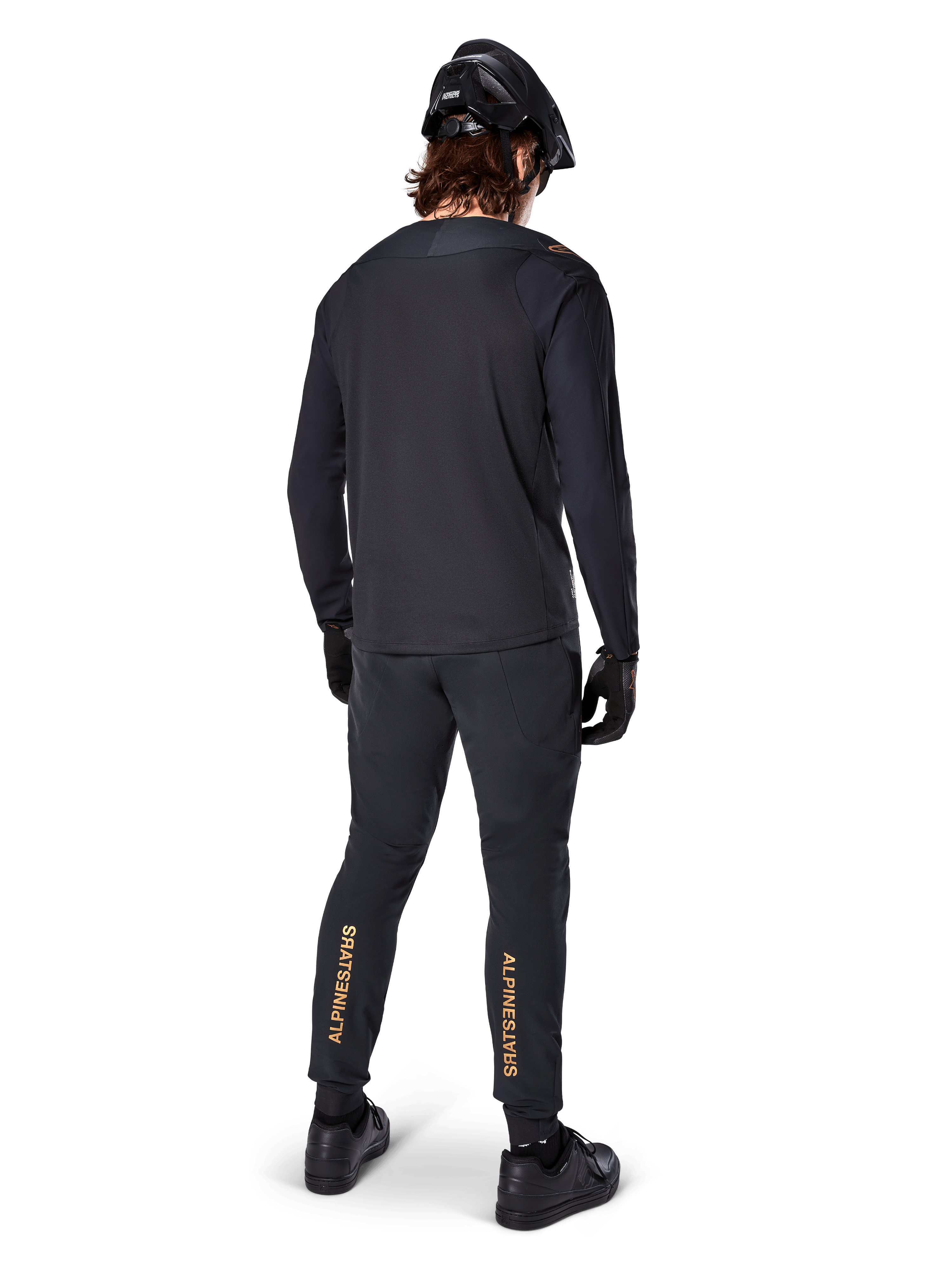 Rear view of a mountain bike rider wearing the Alpinestars A-Aria Polartec Switch Long Sleeve Jersey in Black, paired with black MTB pants featuring tan Alpinestars branding on the calves, black helmet and gloves, professional cycling gear for off-road trails.