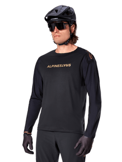 Alpinestars A-Aria Polartec Switch Langarmtrikot, Schwarz mit goldfarbenem Logo, MTB-Mountainbike-Bekleidung, getragen mit schwarzem Helm und Sonnenbrille, mit lasergeschnittenen Belüftungsöffnungen an den Ärmeln und feuchtigkeitsableitendem Polartec-Gewebe für Offroad-Performance