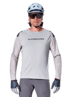 Maillot à manches longues Alpinestars A-Aria Polartec Switch, maillot de VTT gris clair avec perforations laser sur les manches et marquage sur la poitrine, porté avec un casque bleu clair, des lunettes de soleil et des gants gris
