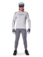 Maillot à manches longues Alpinestars A-Aria Polartec Switch, gris clair, porté par un cycliste de VTT avec pantalon gris foncé, casque bleu clair et lunettes de soleil, avec marquage sur la poitrine et ventilations perforées au laser sur les manches, tenue de cyclisme tout-terrain complète.