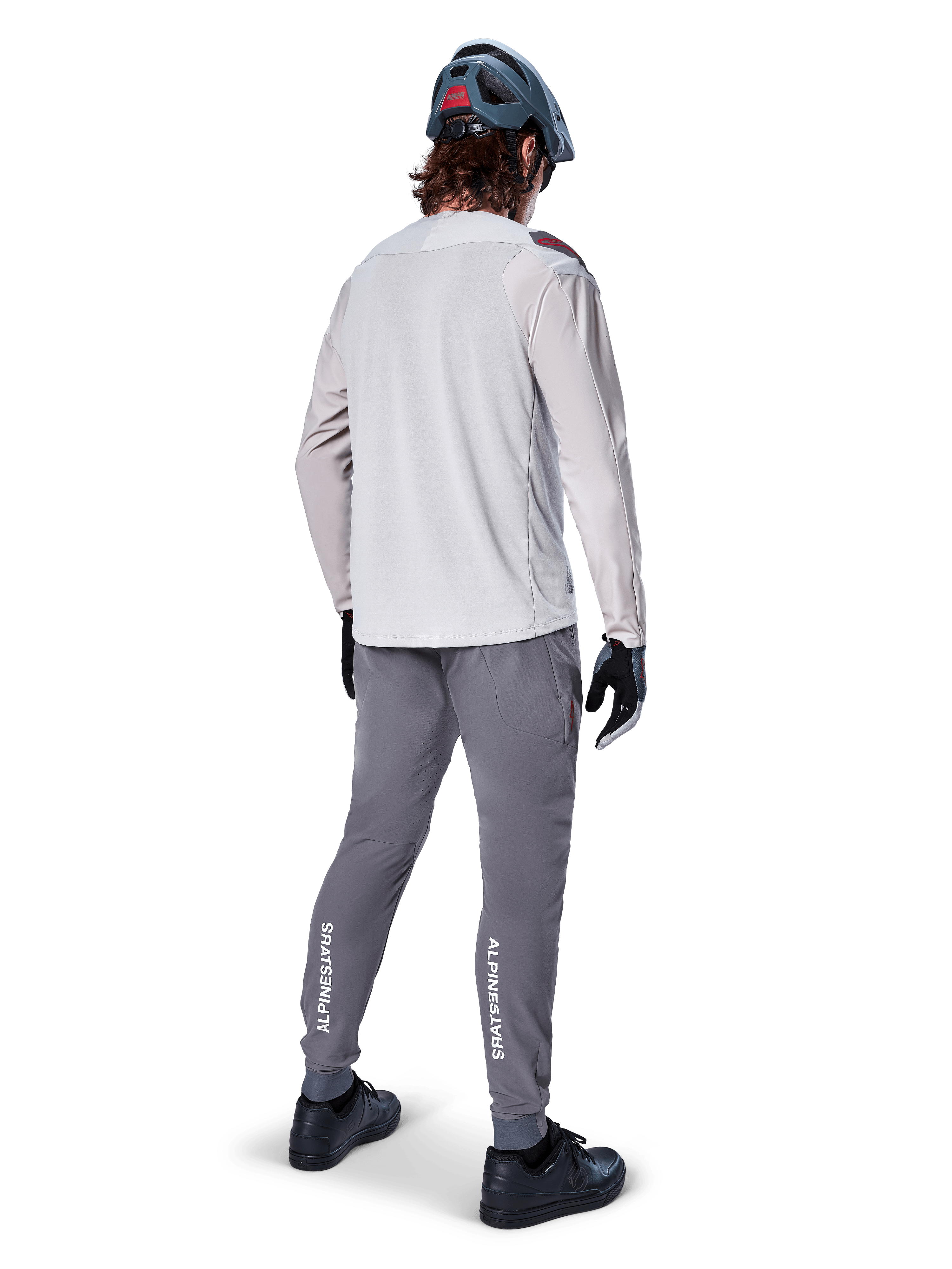 Vue arrière d'un cycliste portant le maillot à manches longues Alpinestars A-Aria Polartec Switch en gris clair, associé à un pantalon de VTT gris foncé avec le logo Alpinestars blanc sur les mollets, des gants noirs et un casque gris.