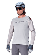 Alpinestars A-Aria Polartec Switch Jersey à manches longues, maillot de cyclisme gris clair avec logo noir sur la poitrine et accent rouge sur l'épaule, manches perforées au laser pour la ventilation, porté avec un casque bleu clair, des lunettes de soleil et un pantalon gris, tenue de VTT haute performance pour le trail