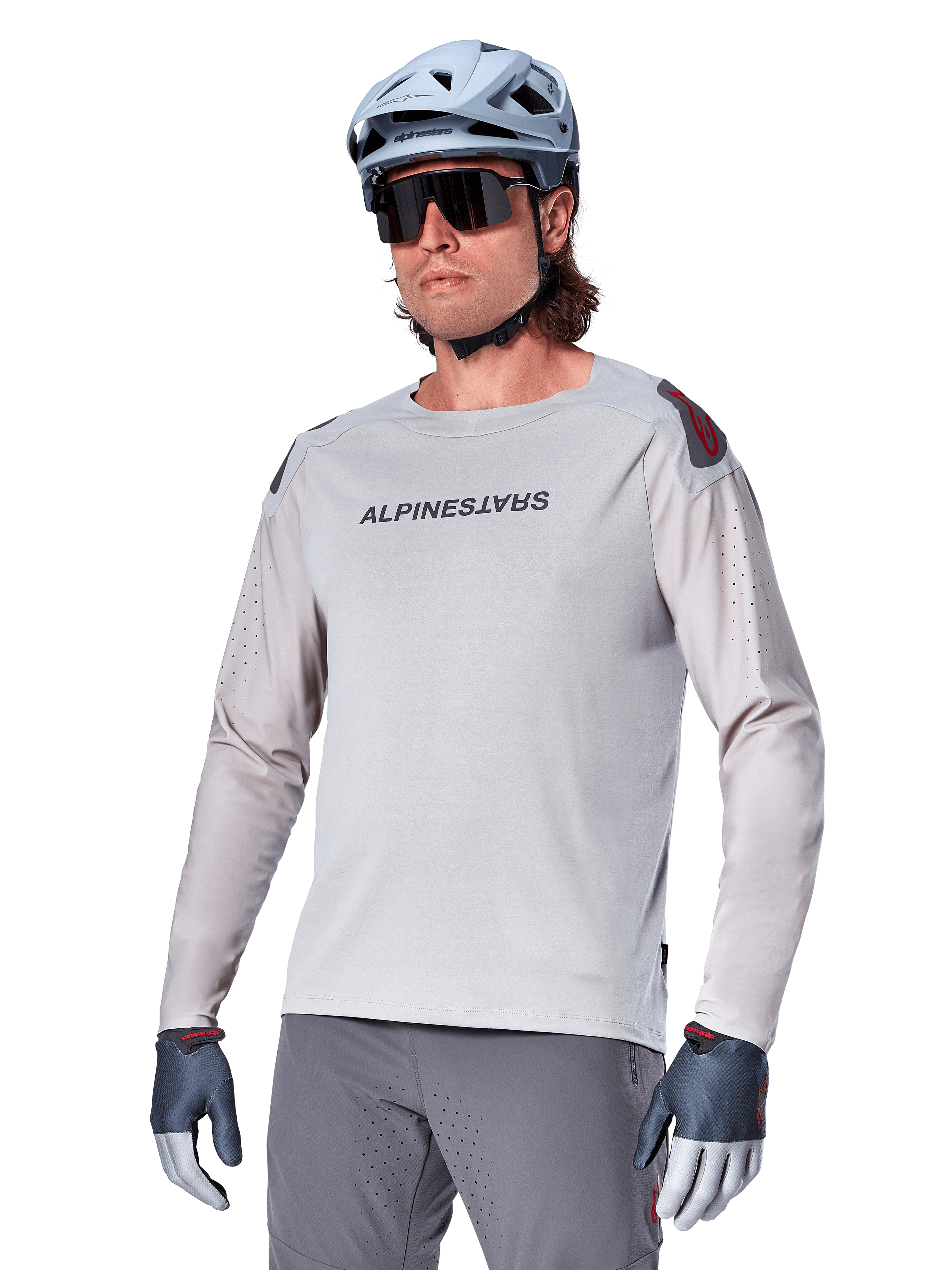 Alpinestars A-Aria Polartec Switch Langarmtrikot, hellgraues Radtrikot mit schwarzem Brustlogo und roten Schulterakzenten, laserperforierte Ärmel für optimale Luftzirkulation, getragen mit hellblauem Helm, Sonnenbrille und grauer Hose, Performance-Mountainbike-Bekleidung für Trailfahrten