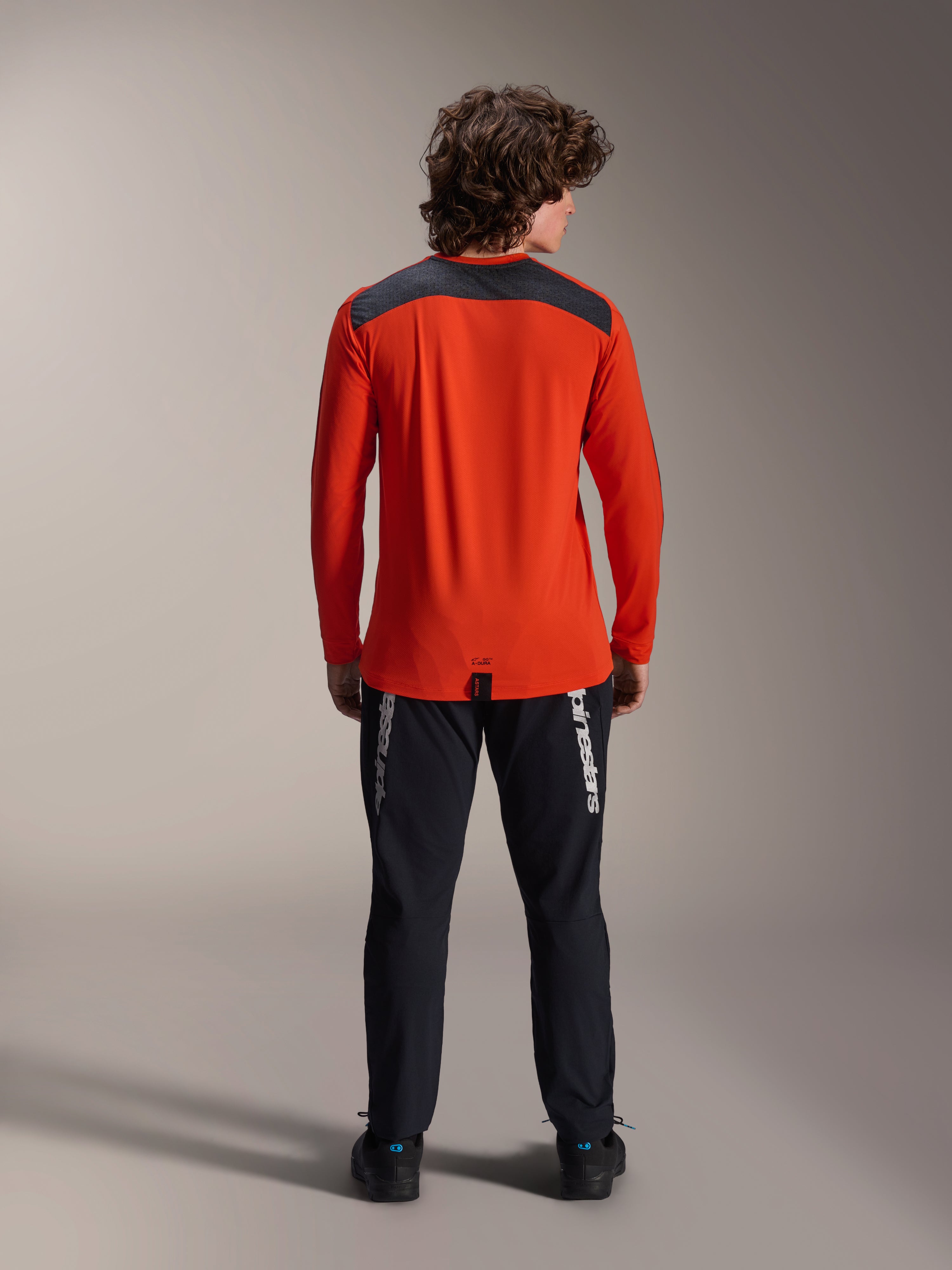 Alpinestars A-Dura Insert Astar Long Sleeve Jersey in Spicy Orange, back view showing charcoal gray shoulder panel, worn with black Alpinestars pants and black shoes, professional studio shot for MTB performance gear
