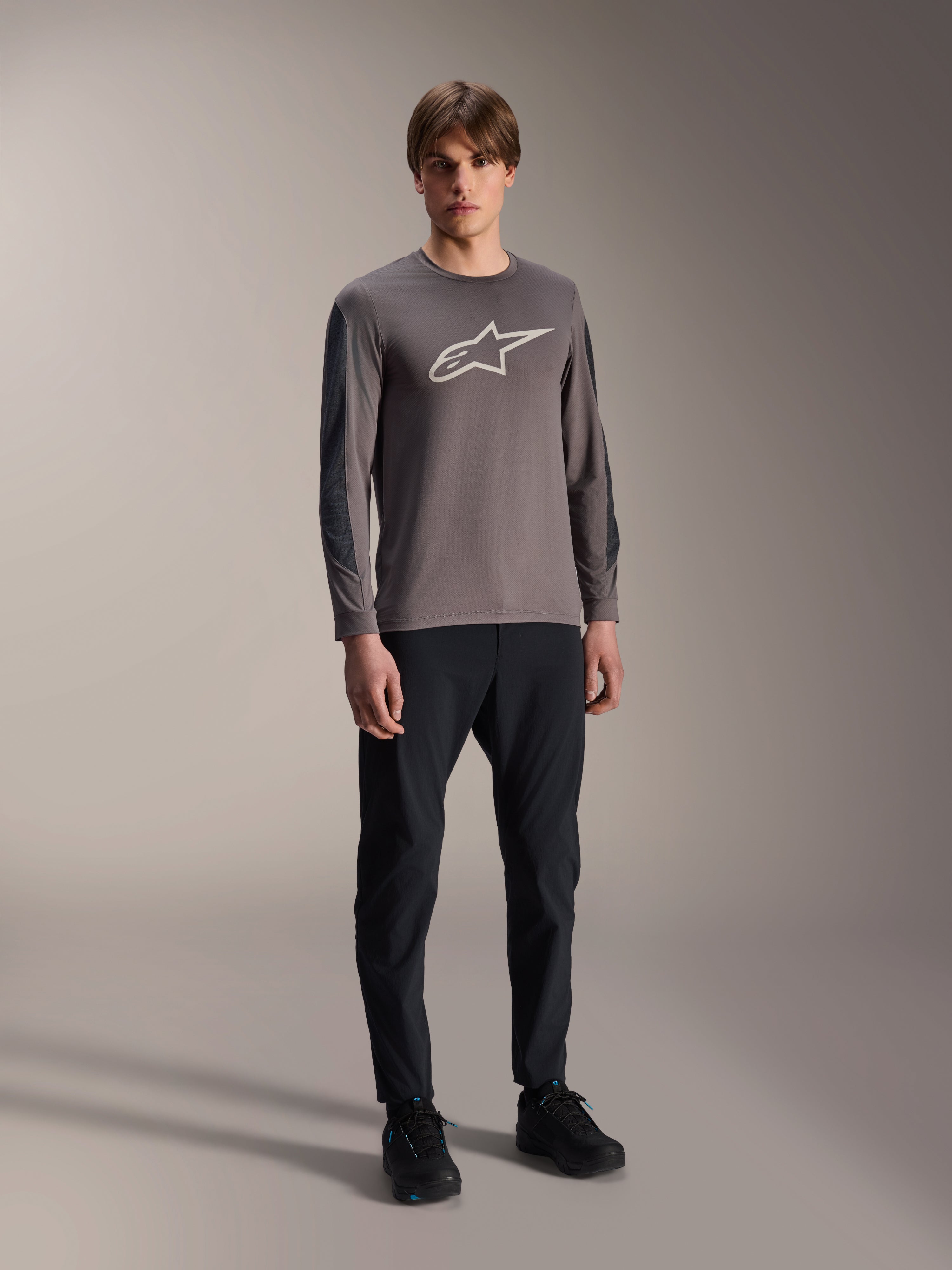 Alpinestars A-Dura Insert Astar Jersey, MTB-shirt met lange mouwen, donkergrijs, met groot wit logo op de borst, gedragen met zwarte technische broek en zwarte schoenen, off-road fietskleding