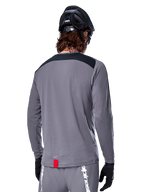 Alpinestars A-Dura Dri Astar Jersey met lange mouwen, donkergrijze MTB-jersey, achteraanzicht met zwart schouderpaneel en klein rood merklabel, gedragen met zwarte mountainbikehelm en handschoenen.