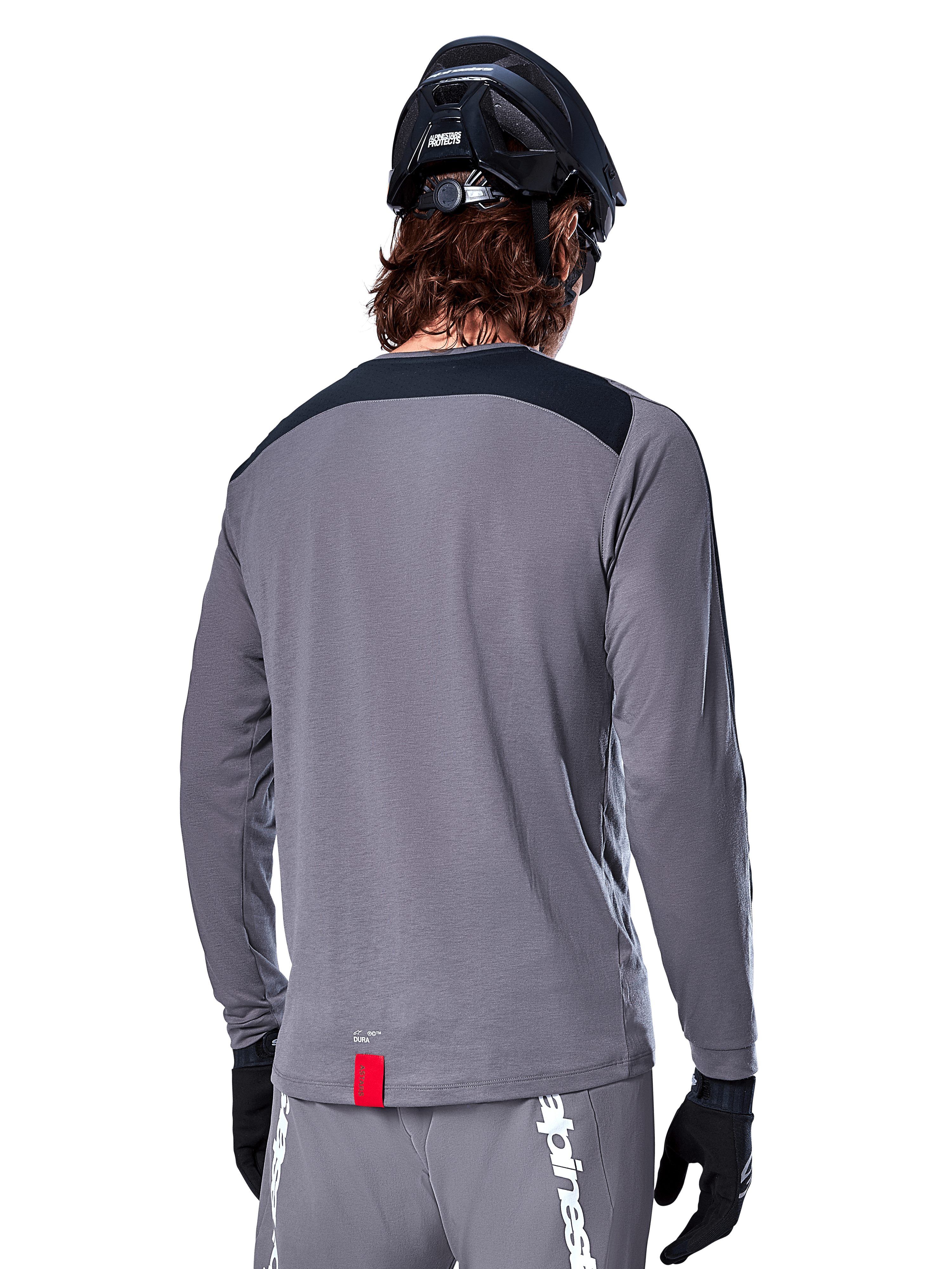 Alpinestars A-Dura Dri Astar Jersey met lange mouwen, donkergrijze MTB-jersey, achteraanzicht met zwart schouderpaneel en klein rood merklabel, gedragen met zwarte mountainbikehelm en handschoenen.