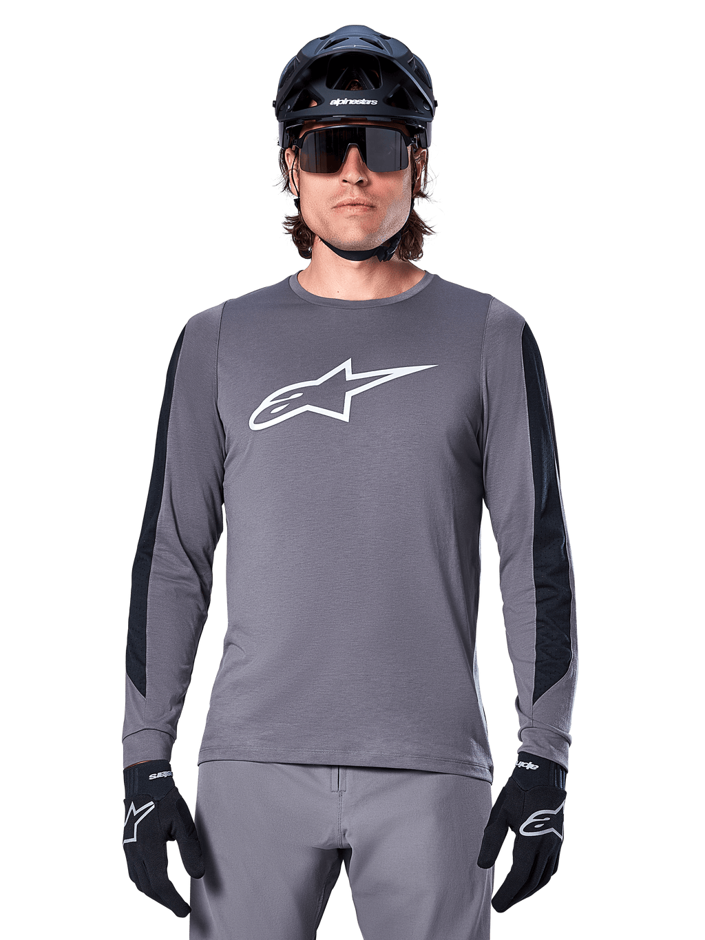 Alpinestars A-Dura Dri Astar Jersey met lange mouwen, donkergrijs, voorzien van een groot wit Astar-logo op de borst en zwarte mesh-panelen op de mouwen, gedragen door een rijder met een zwarte mountainbikehelm en sportzonnebril, MTB-fietskleding