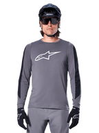 Alpinestars A-Dura Dri Astar Jersey met lange mouwen, donkergrijs, voorzien van een groot wit Astar-logo op de borst en zwarte mesh-panelen op de mouwen, gedragen door een rijder met een zwarte mountainbikehelm en sportzonnebril, MTB-fietskleding